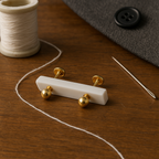 Delicate 18K gold baby earrings with secure design, placed on wooden surface with sewing needle, thread spool, and fabric in background